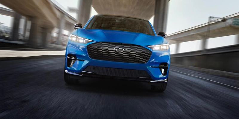 a blue 2025 Mustang Mach-E driving under a bridge