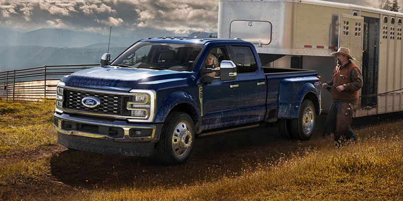 2026 Ford pickup truck towing a large horse trailer through a rugged countryside landscape, with a driver inside and a cowboy standing beside the trailer, highlighting towing capability and durability.