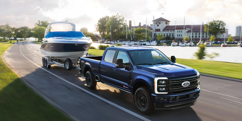 A blue Ford Super Duty F-250 towing a boat on a trailer along a scenic road next to a waterfront, demonstrating its towing power and capability.