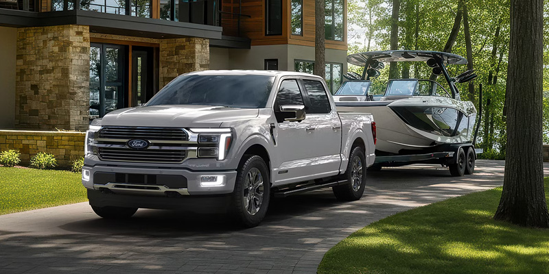 A silver Ford F-150 pickup truck towing a boat on a trailer, parked in front of a modern house surrounded by greenery.